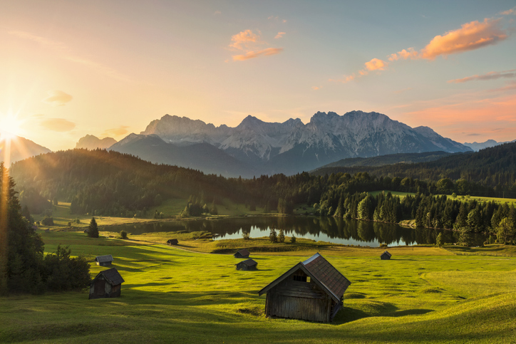 Heuhütte im Vordergrund auf saftiggrüner Wiese. Im Hintergrund eine Bergkette im Sonnenaufgangslicht, die sich teilweise im Bergsee spiegelt.
