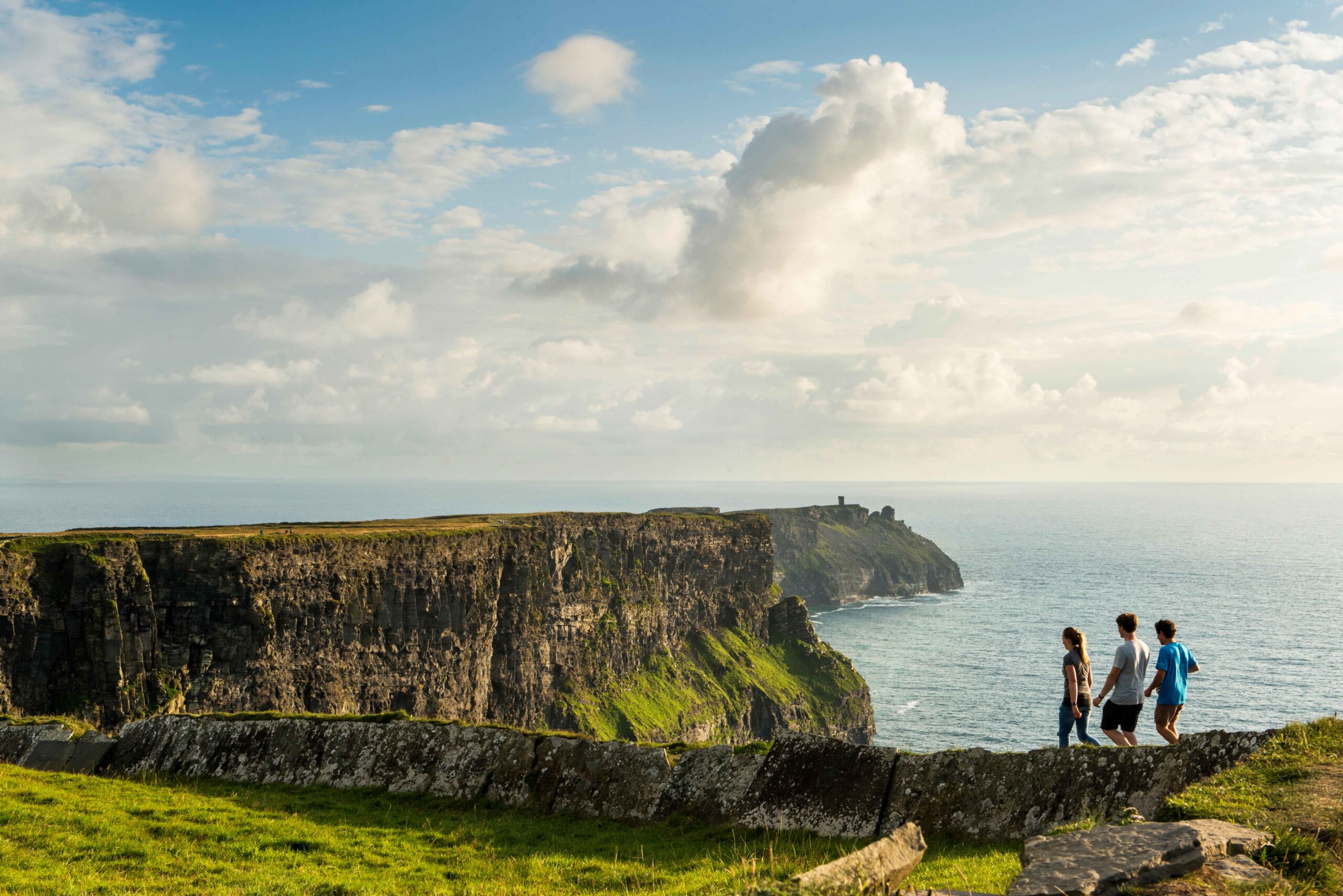 Das Bild zeigt drei Personen, die an den Klippen von Moher in Irland entlanggehen. Im Hintergrund ist der Atlantik mit dem weiten Himmel und einigen Wolken zu sehen. Die Klippen sind hoch und steil und die Szenerie wirkt sehr dramatisch und beeindruckend. Das Licht scheint von vorne zu kommen, wodurch die Steine und das Gras beleuchtet werden. Die Personen sind relativ klein und wirken im Vergleich zur überwältigenden Naturlandschaft sehr bescheiden.