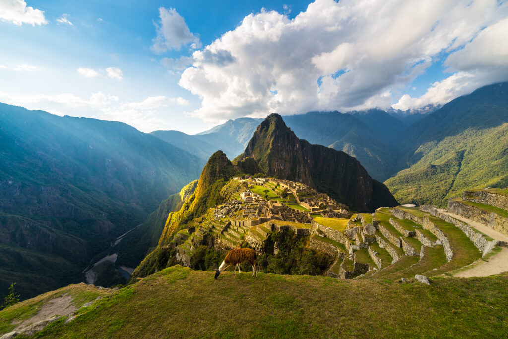 Machu Picchu illuminated by the warm sunset light. Wide angle view from the terraces above with scenic sky and sun burst. Dreamlike travel destination, world wonder. Cusco Region, Peru.