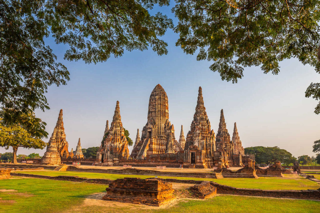 Alte Tempelruinen mit hohen Pagoden in Ayutthaya, Thailand, umgeben von Grünflächen und Bäumen im warmen Sonnenlicht.