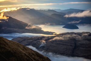 Blick auf Lake Wanaka und die Berglandschaft von Treble Cone in Neuseeland, mit grünen Hügeln und klarer Sicht unter sonnigem Himmel.