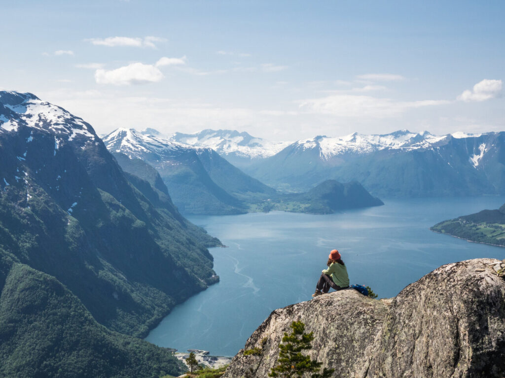 Frau sitzt auf Felsen und schaut auf Fjordpanorama mit schneebedeckten Bergen