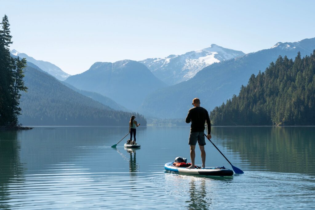 Zwei Personen stehen auf Stand-up-Paddle-Boards auf einem ruhigen See vor bewaldeten Bergen mit schneebedeckten Gipfeln