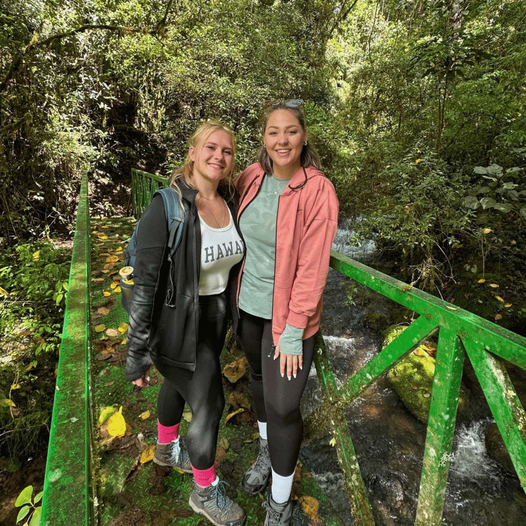 Anika und Isa stehen auf einer grünen Metallbrücke über einem Bach, umgeben von dichtem Wald und Moos.
