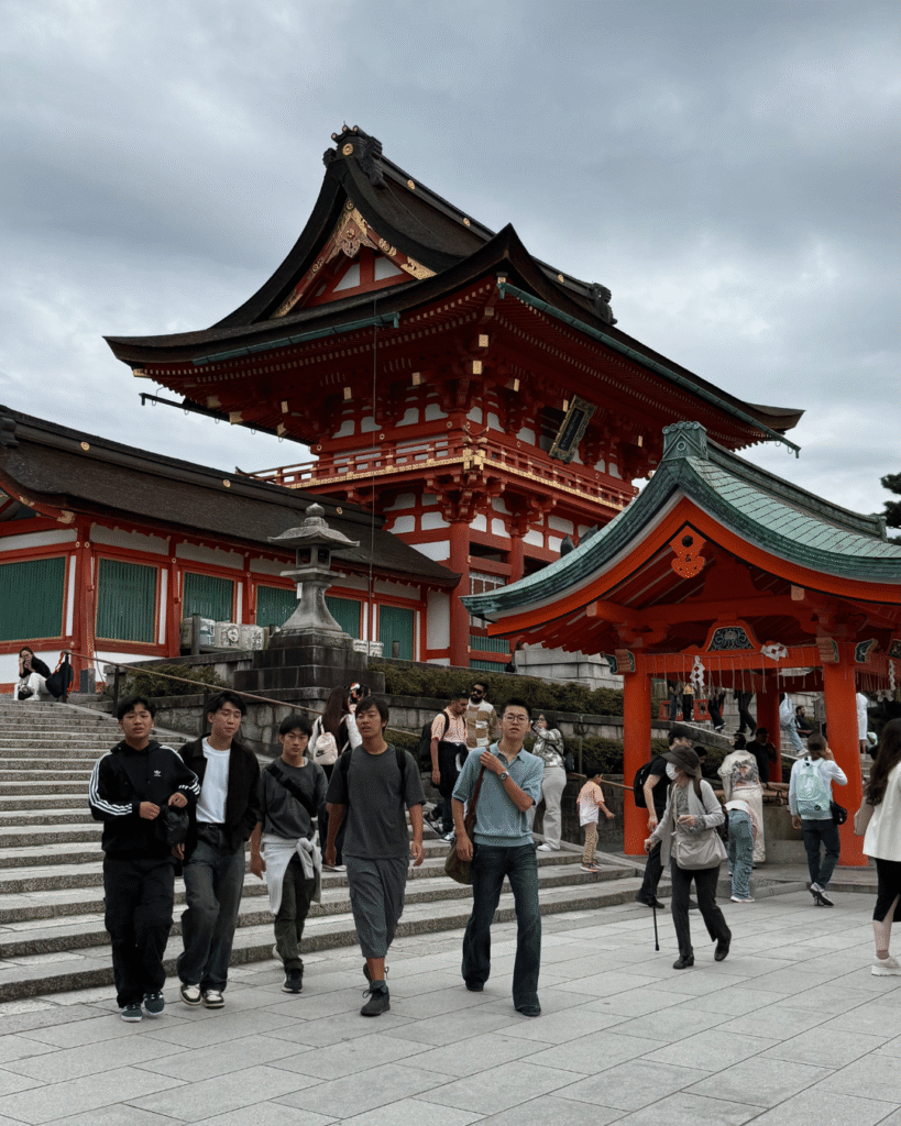 Besucher vor dem Fushimi Inari-Taisha Schrein in Kyoto mit rotem Torii und traditioneller Architektur unter bewölktem Himmel.
