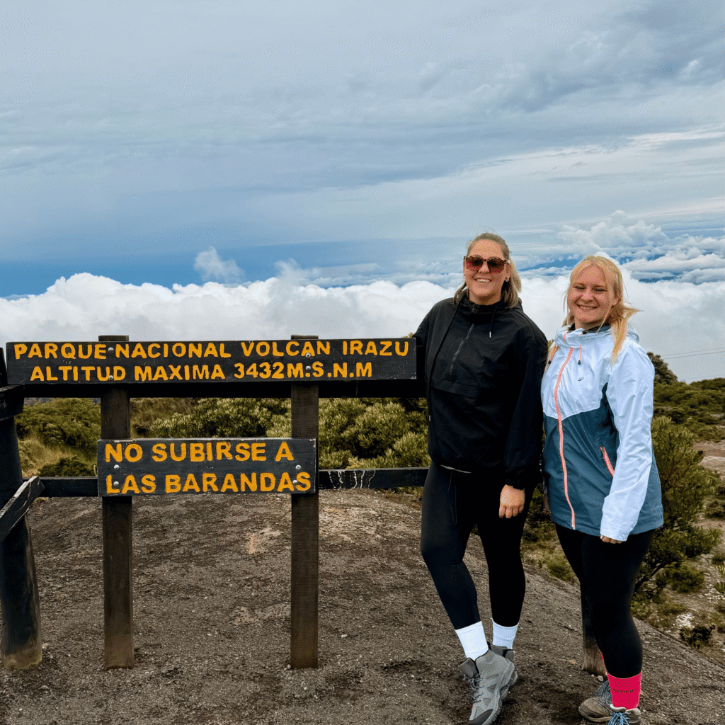 Zwei Frauen posieren neben einem Holzschild mit der Aufschrift „Parque Nacional Volcán Irazú, Altitud Máxima 3432 m.s.n.m.“ vor Wolkenlandschaft.