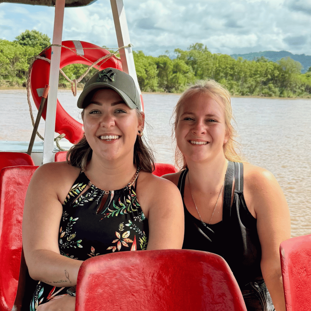 Zwei Frauen itzen auf roten Kunststoffsitzen in einem offenen Boot auf einem Fluss, im Hintergrund grüne Ufervegetation und Berge.