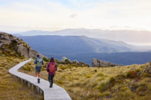 Zwei Wanderer auf dem Hump Ridge Track in Fiordland, Neuseeland, auf Holzsteg durch alpine Landschaft mit Blick auf Berge und Täler bei Sonnenaufgang.