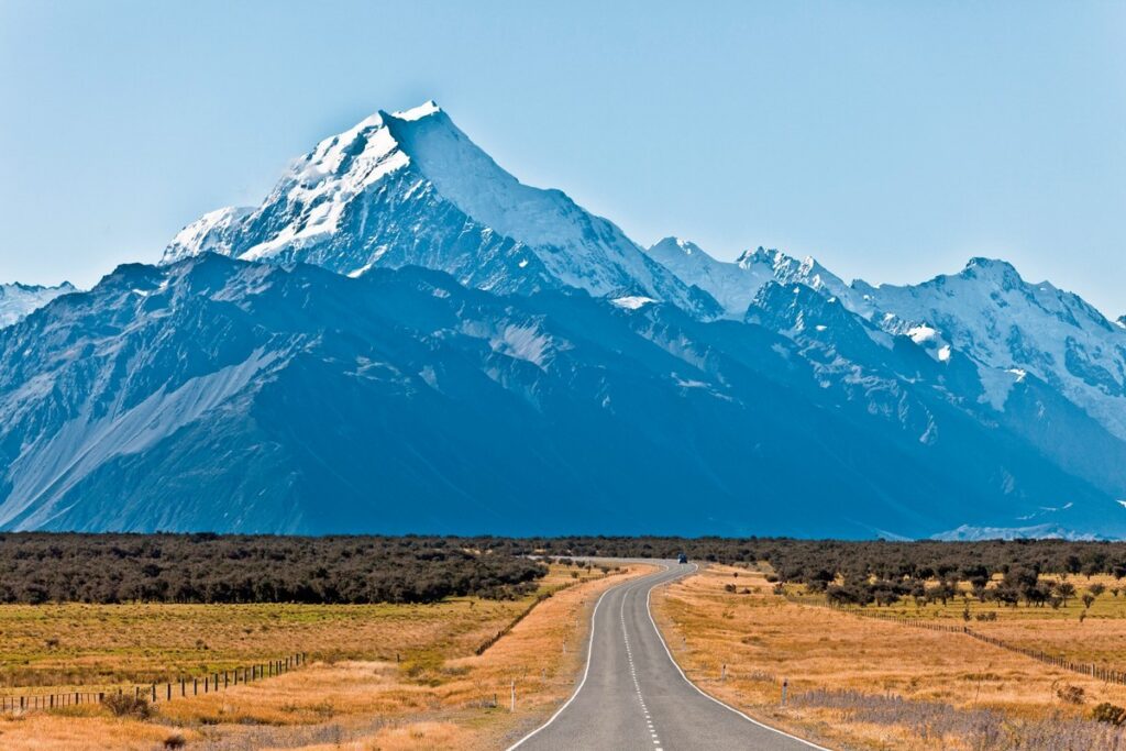 Straße führt durch weite Ebene zum schneebedeckten Aoraki/Mount Cook in Neuseeland unter klarem blauem Himmel