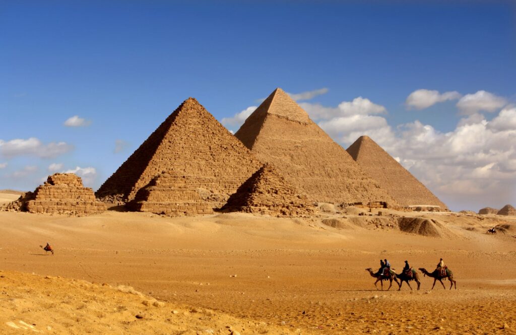 Die Pyramiden von Gizeh in der ägyptischen Wüste unter blauem Himmel, mit drei Kamelen und Reitern im Vordergrund