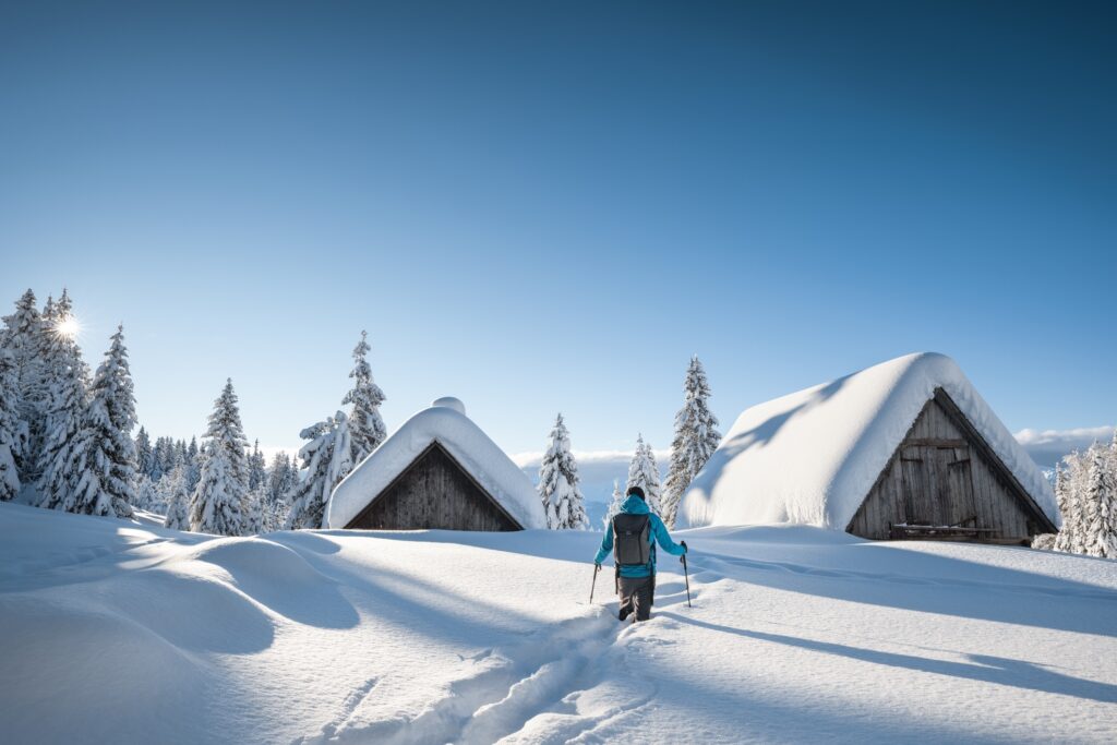 Wanderer geht mit Stöcken durch tiefen Schnee zu drei Hütten mit schneebedeckten Dächern, umgeben von verschneiten Bäumen.