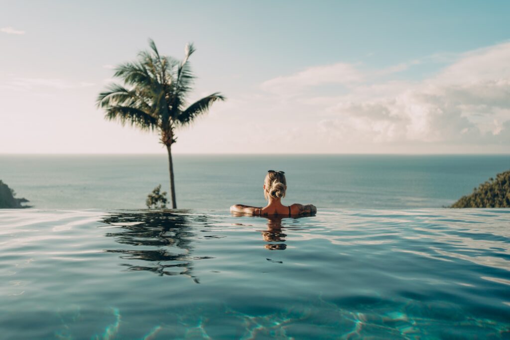 Frau entspannt im Infinity-Pool mit Blick auf das Meer, Palmen und bewaldete Hügel unter klarem Himmel.