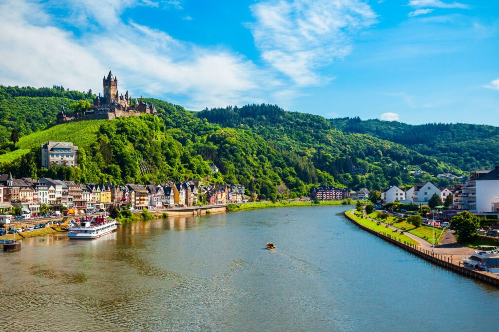 Blick auf die Mosel mit Weinbergen, der Burg Cochem auf einem Hügel und farbenfrohen Häusern am Flussufer unter blauem Himmel.