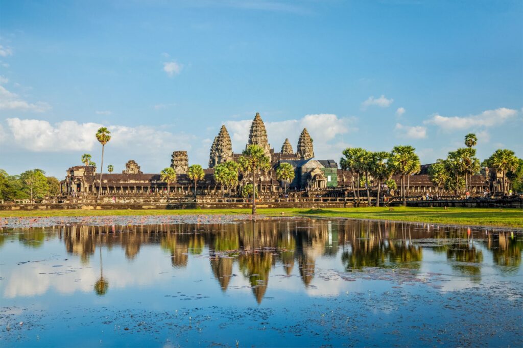 Angkor Wat, das berühmte Tempelensemble in Kambodscha, spiegelt sich im ruhigen Wasser, umgeben von Palmen unter klarem blauem Himmel.