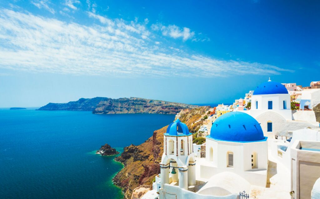 Weiße Kirche mit blauen Kuppeln in Oia auf Santorin, Griechenland, über der Ägäis mit weitem Blick auf das tiefblaue Meer und die Küste.