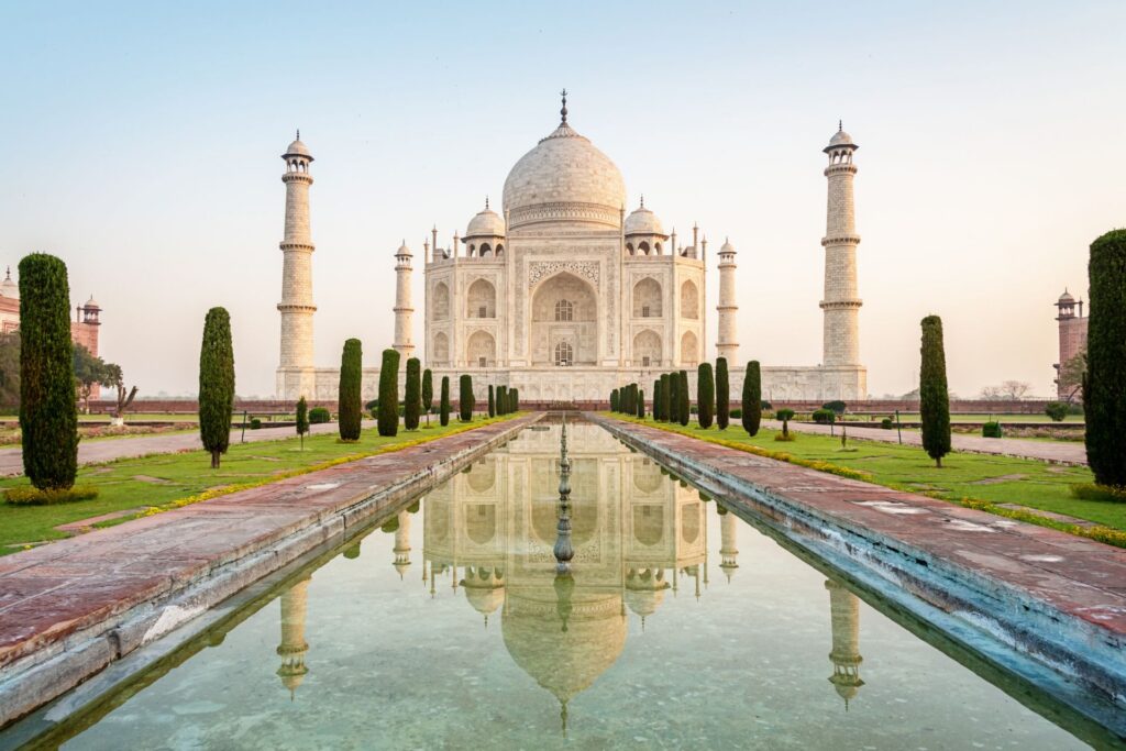 Das Taj Mahal in Agra, Indien, bei Sonnenaufgang. Die weiße Marmorkuppel spiegelt sich im Wasserbecken, umgeben von gepflegten Gärten und Minaretten.