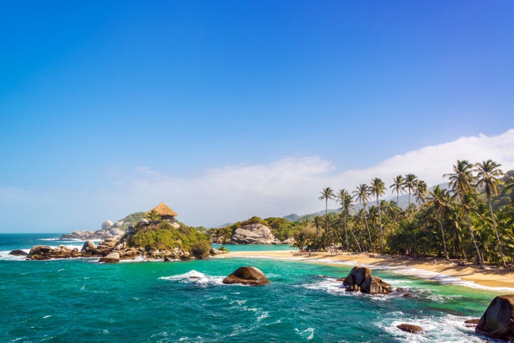 Tropischer Strand im Tayrona-Nationalpark, Kolumbien: türkisblaues Karibikwasser, Palmen und Felsen unter klarem, sonnigem Himmel.