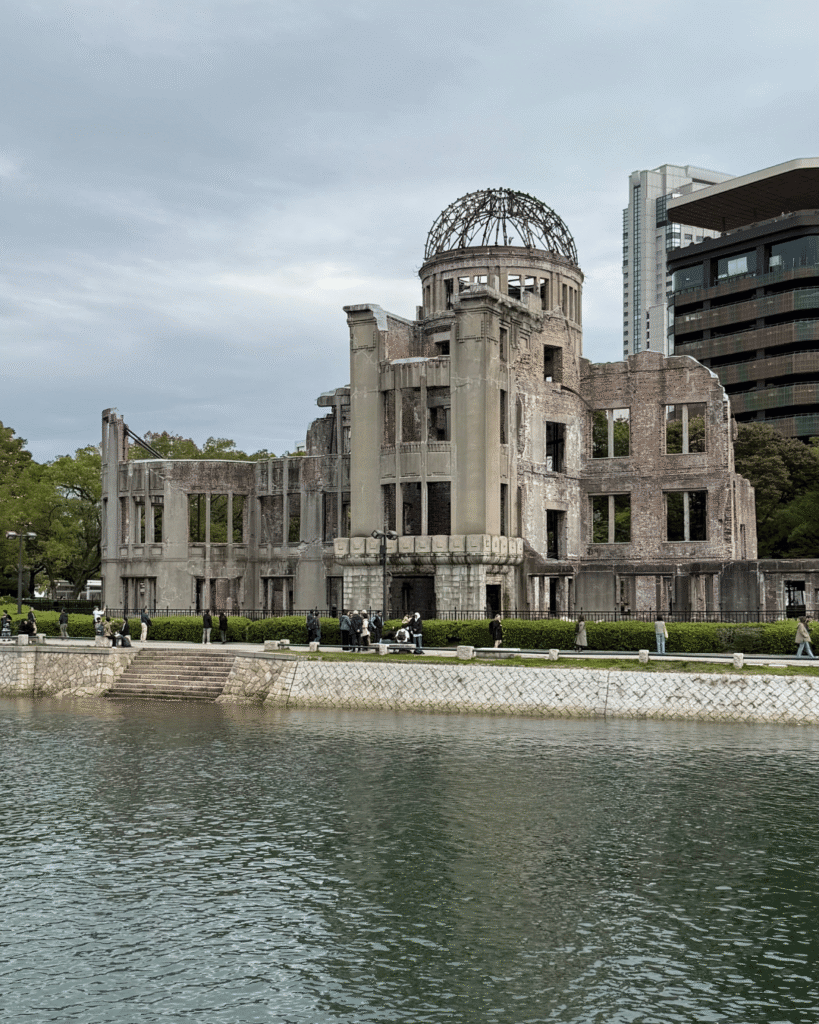 Atombomben-Dom in Hiroshima am Flussufer, historische Ruine des Friedensdenkmals mit grünen Bäumen und modernen Gebäuden im Hintergrund unter bewölktem Himmel.