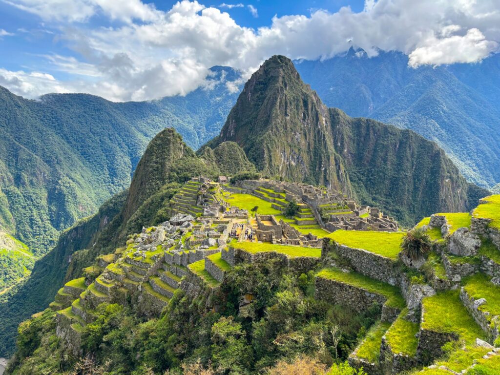 Machu Picchu in Peru unter blauem Himmel mit dramatischen Wolken: antike Terrassen und Ruinen eingebettet in grüne Andenlandschaft am Morgen.