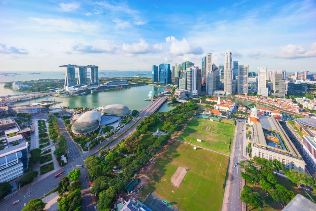 Luftaufnahme von Singapur mit Marina Bay Sands, modernen Wolkenkratzern, grünen Parks und dem Hafen unter blauem Himmel.
