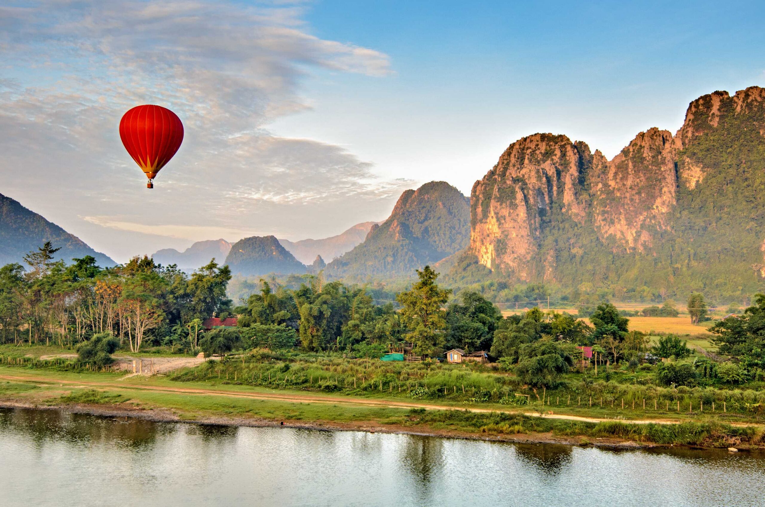 Vang Vieng, Laos - Heißluftballon schwebt über der grünen Landschaft mit grün behangenen Bergen im Hintergrund und einem Fluss im Vordergrund