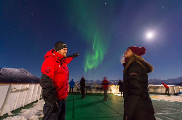 Nordlichter über dem Deck eines Hurtigruten Schiffs in Norwegen, Reisende beobachten das Polarlicht unter klarem Nachthimmel mit Mond und verschneiten Bergen