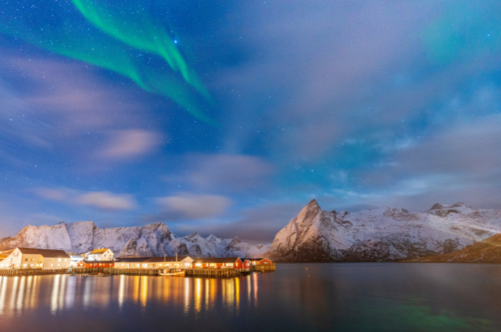 Nordlichter über verschneiten Bergen und roten Fischerhütten auf den Lofoten in Norwegen bei Nacht, Spiegelung im ruhigen Wasser