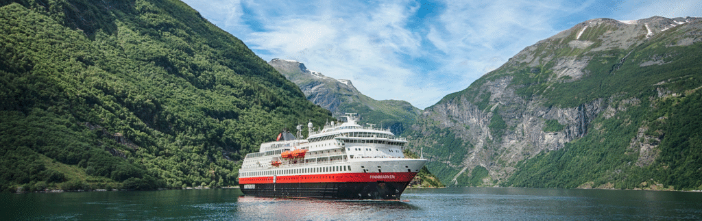 Kreuzfahrtschiff im Geirangerfjord in Norwegen, umgeben von grünen Bergen und klarem Wasser unter blauem Himmel – Hurtigruten Reise. © Foto: Agurtxane Concellon