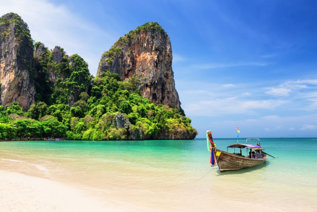 Tropischer Strand mit türkisfarbenem Wasser und einem traditionellen Holzboot im seichten Meer. Dahinter ragen grüne, steile Felsen in den blauen Himmel und prägen die malerische Küstenkulisse.