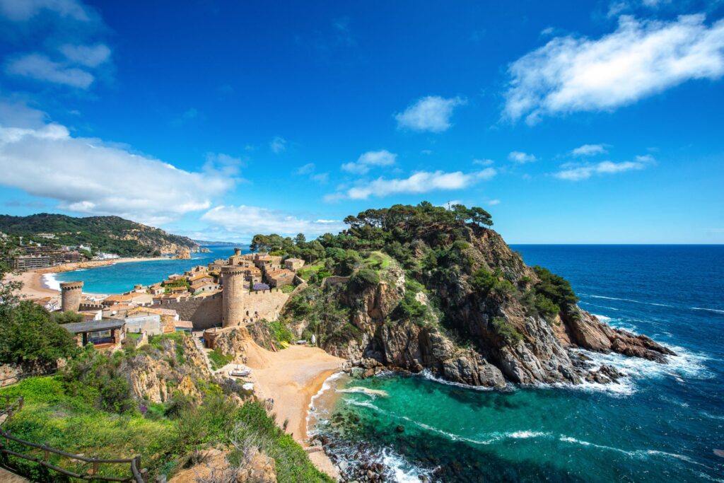 Küstenort mit historischen Mauern liegt an einer türkisfarbenen Bucht, umgeben von felsigen Klippen, Pinien und dem tiefblauen Meer unter blauem Himmel.