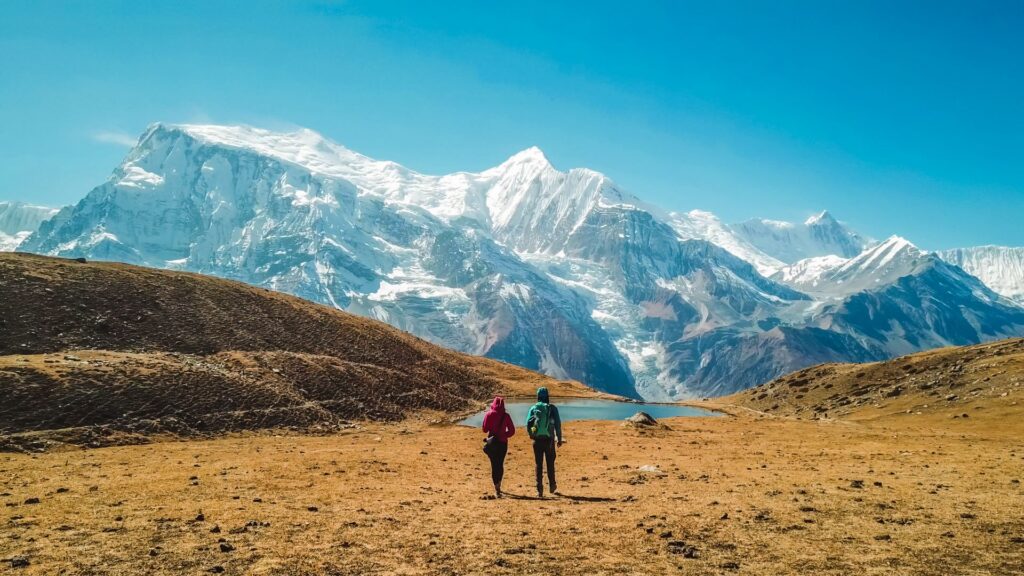 Zwei Personen wandern über eine weite Hochebene auf ein schneebedecktes Bergmassiv zu, das unter strahlend blauem Himmel beeindruckend aufragt.
