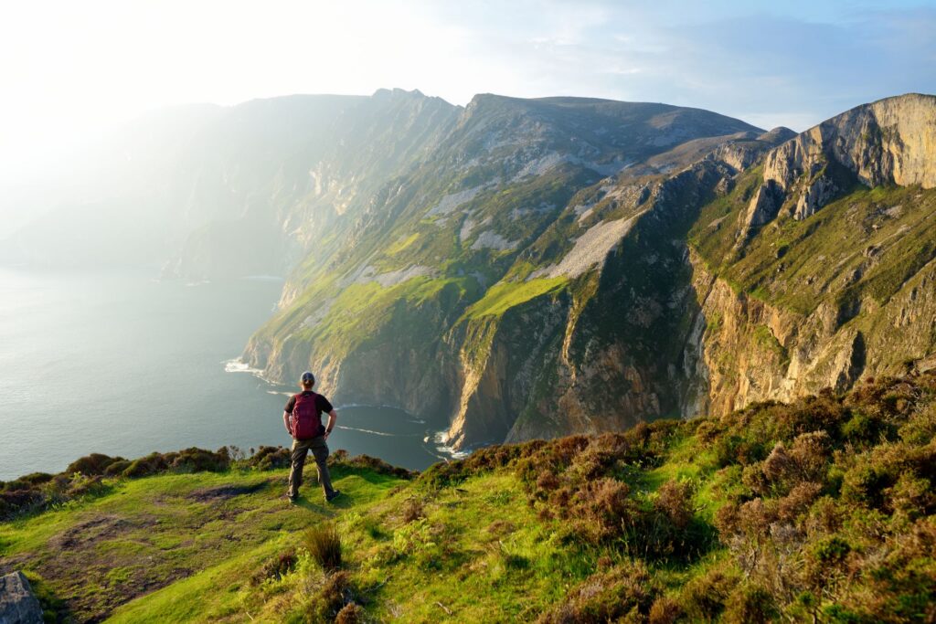 Ein Wanderer steht auf einer grünen Klippe mit Blick auf steil abfallende Felsen und das ruhige Meer, während warmes Licht die Landschaft einhüllt.