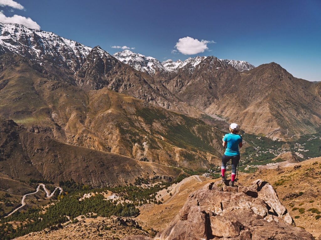 Eine Person steht auf einem Felsen und blickt auf ein weites Gebirgspanorama mit schneebedeckten Gipfeln und Tälern unter klarem, blauem Himmel.