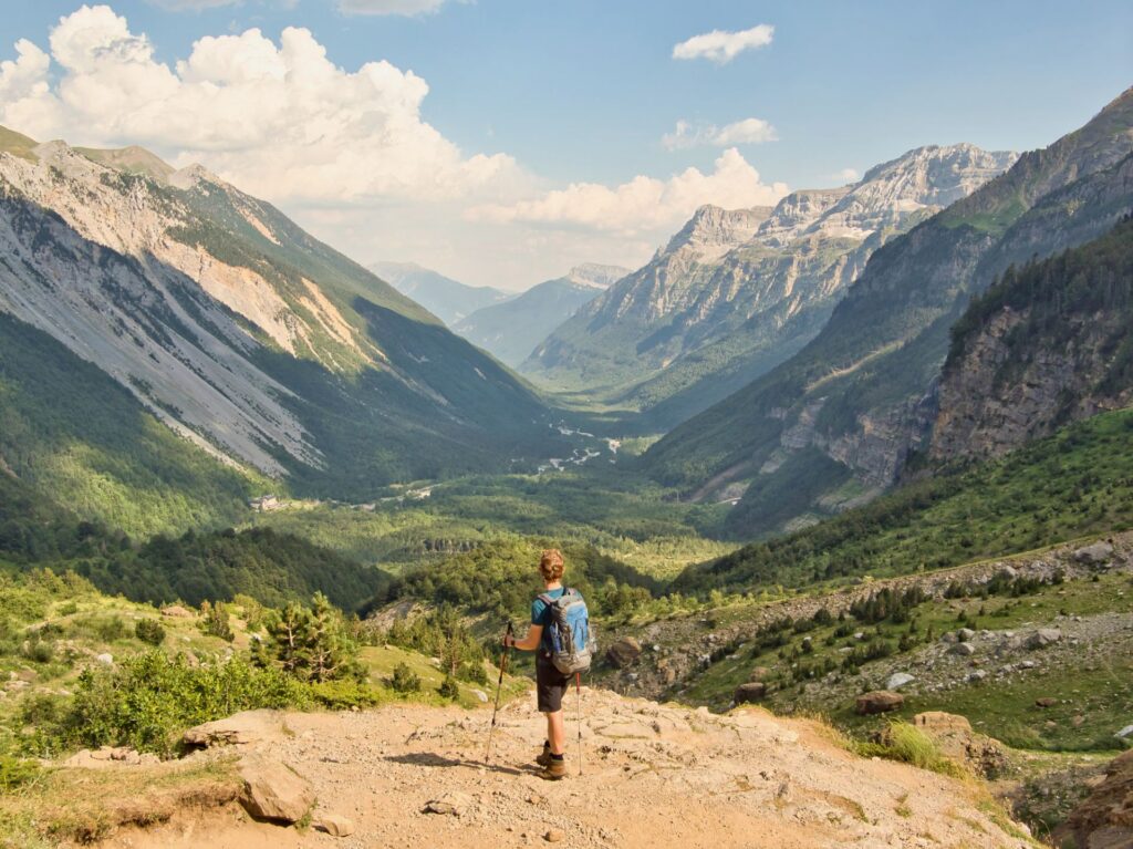 Person wandert auf einem Bergpfad und blickt über ein weites, grünes Tal, umgeben von steilen Felswänden und sonniger, klarer Berglandschaft.