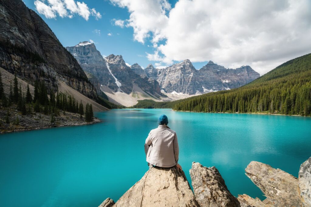 Man sitzt auf Stein und blickt auf blauen See mit Bergkette im Hintergrund.