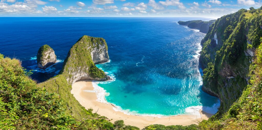 Atemberaubende Aussicht auf eine türkisfarbene Bucht mit weißem Strand, umgeben von steilen Felsen und tiefblauem Meer unter klarem Himmel.