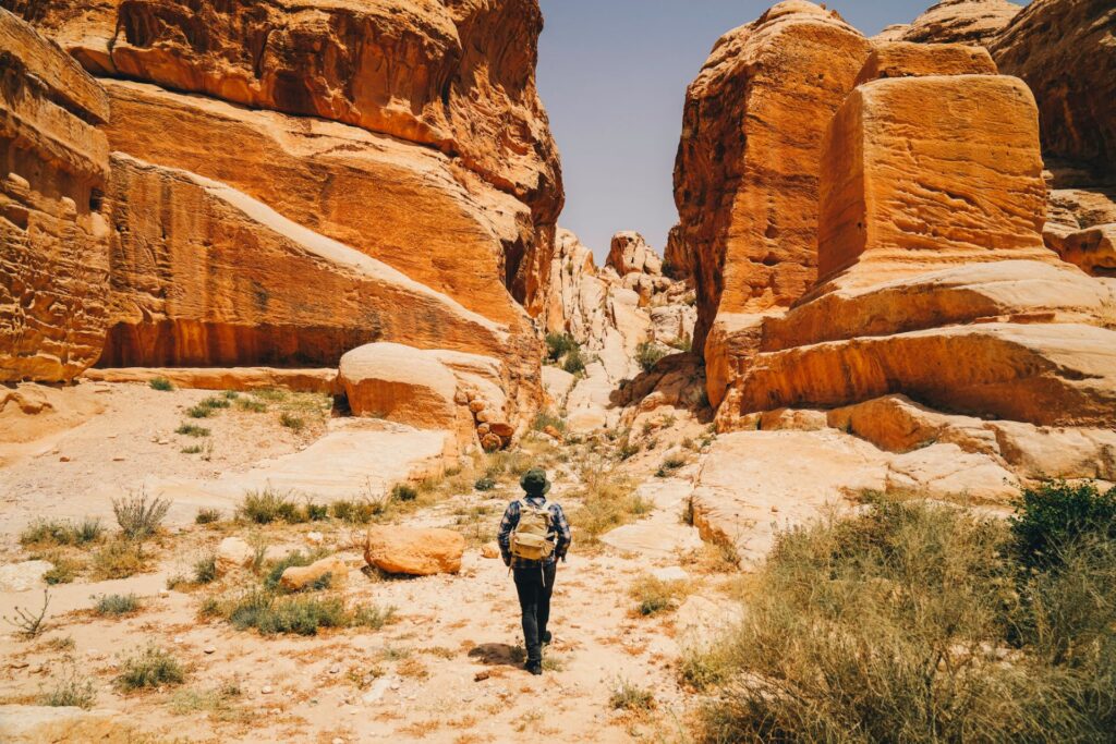 Eine Person wandert mit Rucksack durch eine enge, sonnige Felsenschlucht, umgeben von intensiven Orange- und Sandtönen der hohen Gesteinswände.