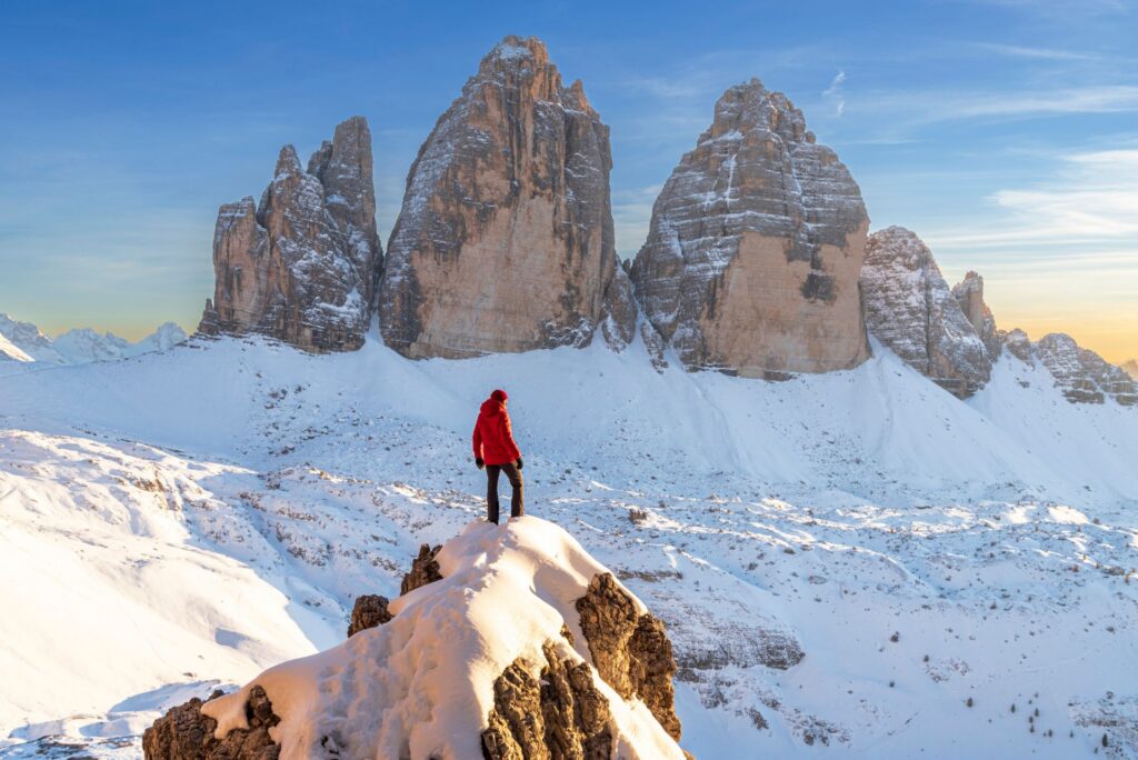 Eine Person in roter Jacke steht auf einem verschneiten Felsvorsprung und blickt auf die markanten, sonnenbeschienenen Gipfel einer winterlichen Bergkette (Drei Zinnen, Südtirol).