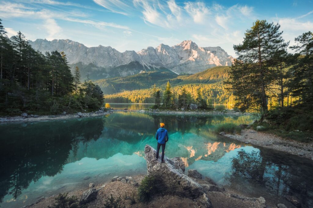Ein Mann genießt die atemberaubende Morgenlandschaft an einem wunderschönen See in den bayerischen Alpen. Das türkisfarbene Wasser spiegelt die Bergkette und die sanften Wolken wider.