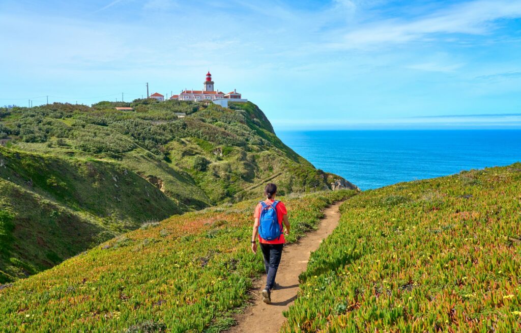 Person wandert auf Küstenpfad über grüne Hügel in Richtung Leuchtturm, mit weitem Blick auf das Meer und einem klaren, sonnigen Himmel.