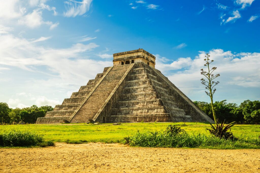 Steinstufenpyramide erhebt sich unter blauem Himmel auf einer weiten, sonnigen Wiese, umgeben von Vegetation und ruhiger Landschaft.