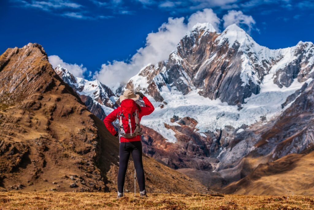 Eine wandernde Person mit roter Jacke blickt auf ein imposantes Bergmassiv mit Schnee und Felsen, das sich unter blauem Himmel beeindruckend erhebt.