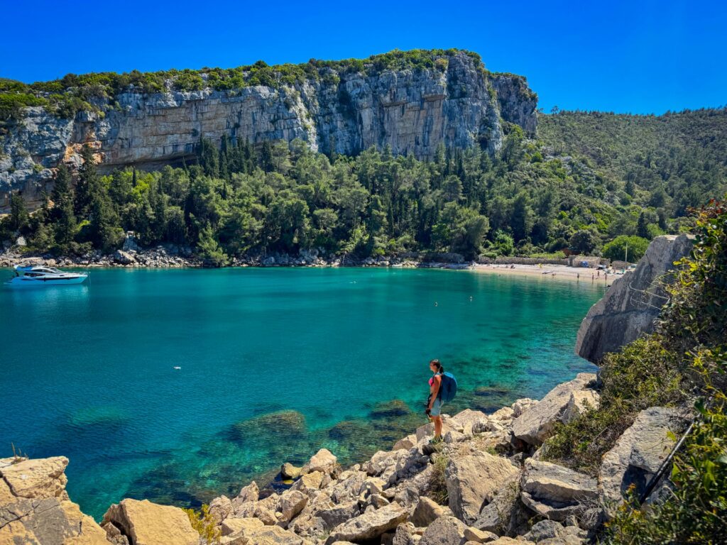 Frau steht am felsigen Ufer und blickt auf eine ruhige türkisfarbene Bucht