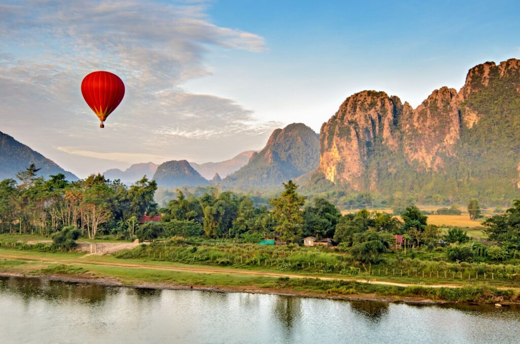 Ein roter Heißluftballon schwebt über einer grünen Flusslandschaft vor hohen Felsbergen, die im warmen Morgenlicht leuchten.