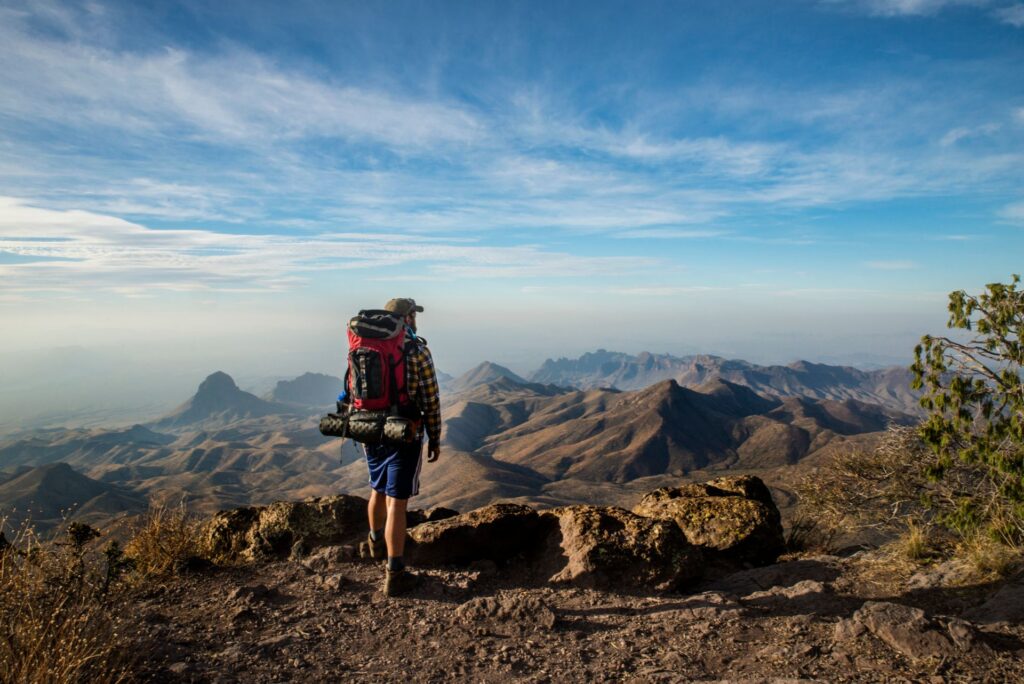 Eine Person mit Rucksack steht an einem Felsvorsprung und blickt über eine weite Berglandschaft unter hellem, leicht bewölktem Himmel.