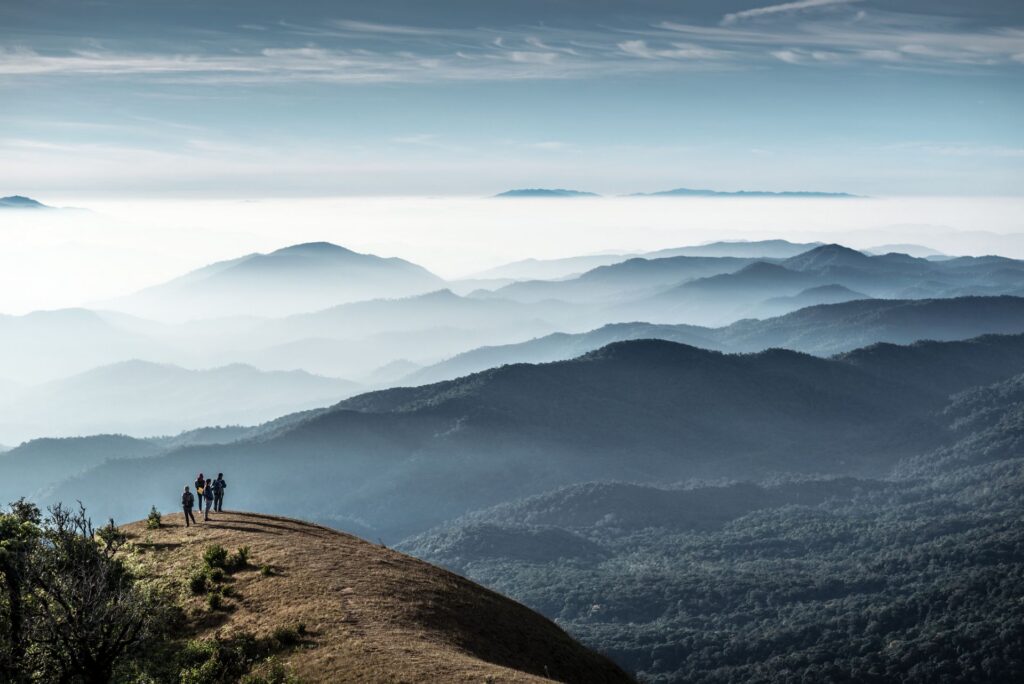 Weitläufige Berglandschaft mit sanften, übereinanderliegenden Hügelketten unter einem hellen Himmel. Eine kleine Gruppe von Menschen steht auf einem Hügel und blickt über das Nebelmeer.