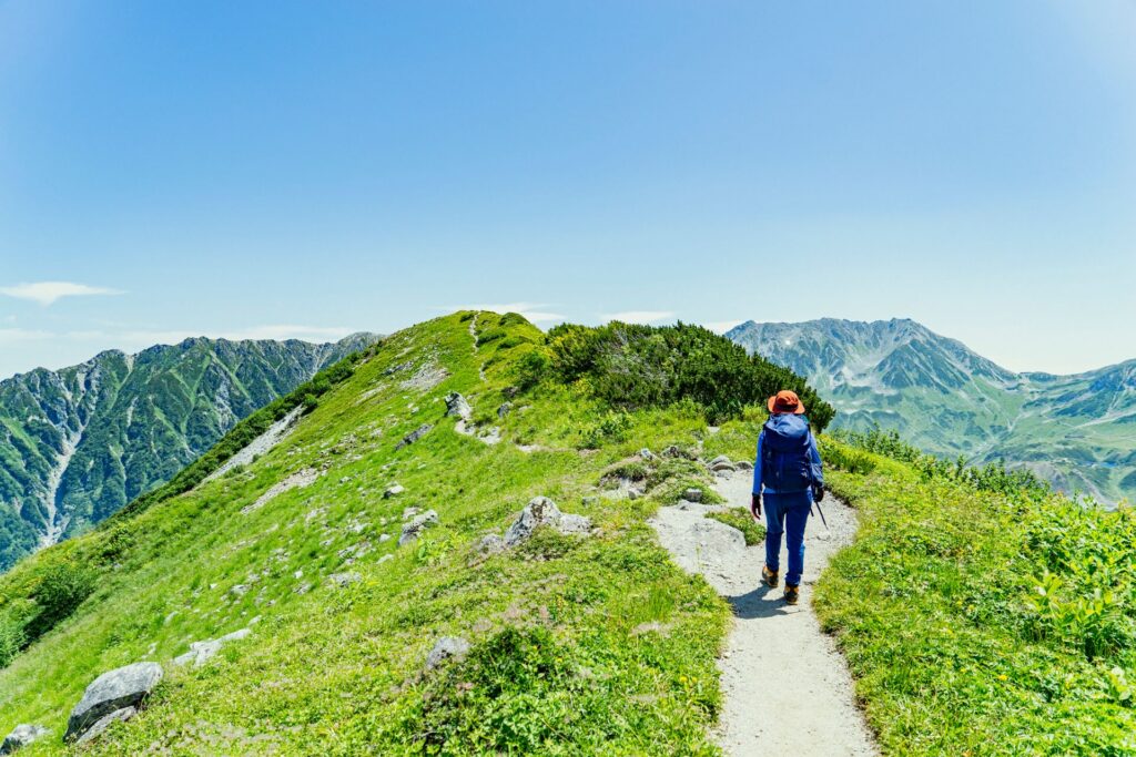 Eine wandernde Person auf einem schmalen Bergpfad, umgeben von grünen Hügeln und klarer Sicht auf umliegende Gebirgsketten bei sonnigem Wetter.