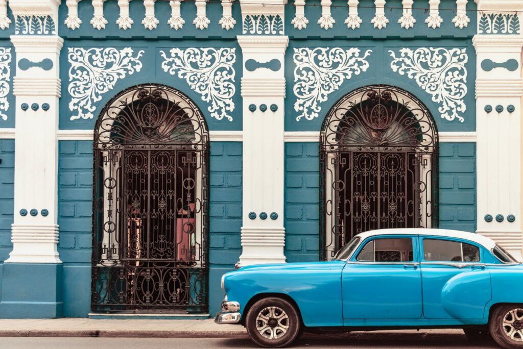 Vor einer reich verzierten, türkis-weißen Fassade steht ein klassischer blauer Oldtimer, der die historische Atmosphäre der Straße unterstreicht.