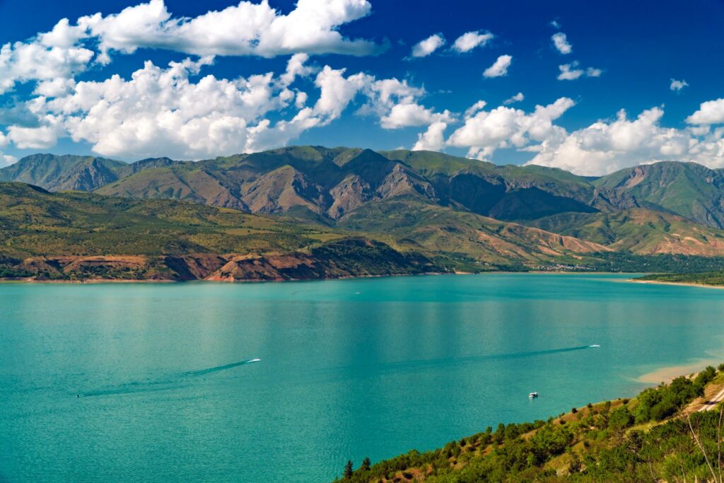 Ein türkisfarbener See liegt vor einer weitläufigen Bergkette unter blauem Himmel mit weißen Wolken, während Boote feine Spuren über das Wasser ziehen.
