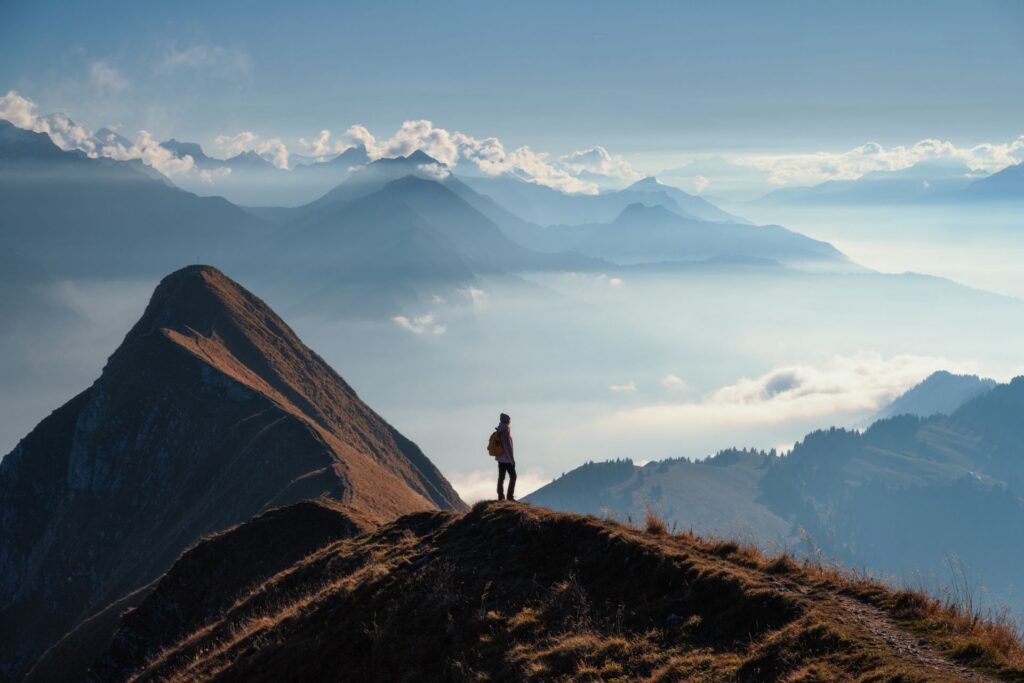 Eine einzelne Person steht auf einem Bergrücken und blickt über nebelverhangene Täler und markante Gipfel, die im weichen Morgenlicht sichtbar werden.